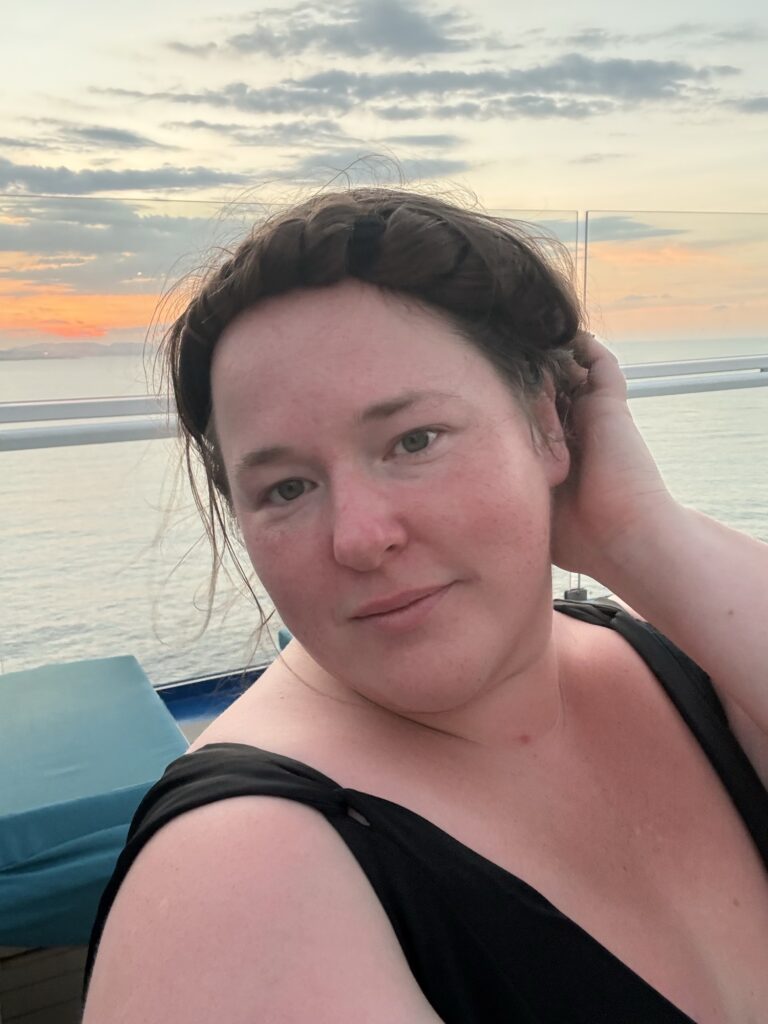 Bobbie JoEle, travel writer at My Vacationer standing on a cruise ship upper deck with the sunset behind her and the wind in her hair